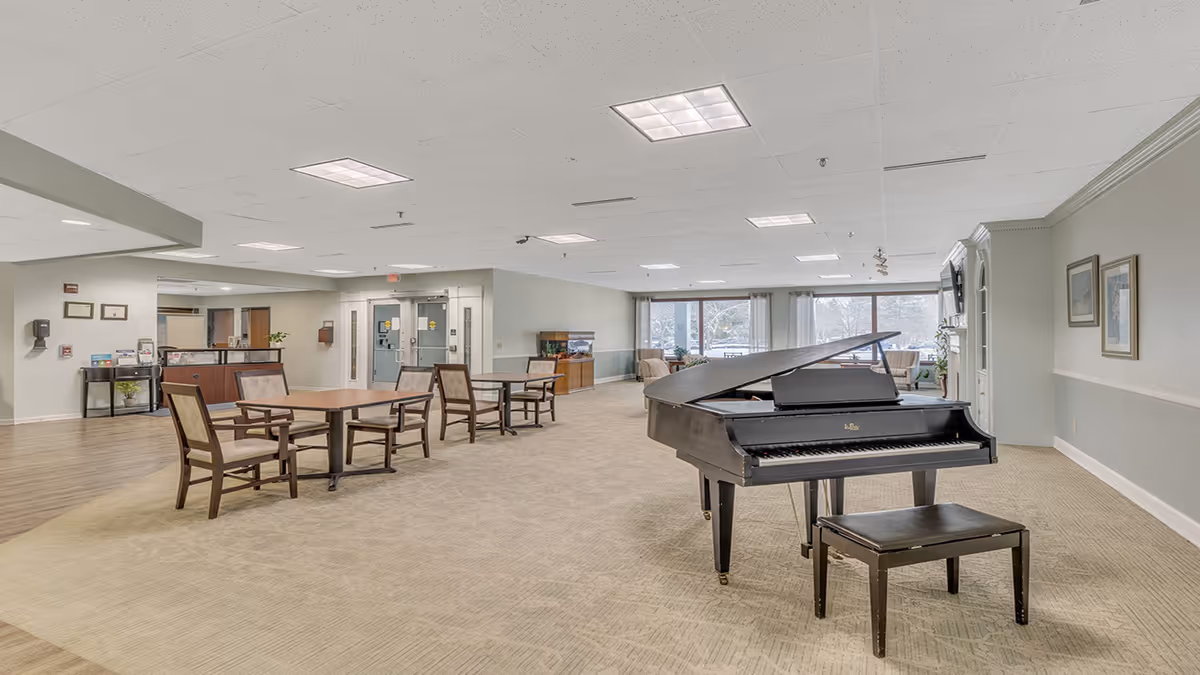 A spacious senior living facility common area with a grand piano and piano bench in the foreground. The room has several tables and chairs, large windows with curtains letting in natural light, and a cozy seating area with armchairs near the windows. The walls are light-colored, and the ceiling has recessed lighting.