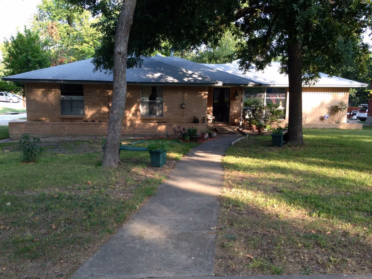Single-story brick building with a gray shingled roof, surrounded by green grass and trees. A concrete walkway leads up to the front door, which is partially shaded by trees. There are potted plants near the entrance and windows with curtains.