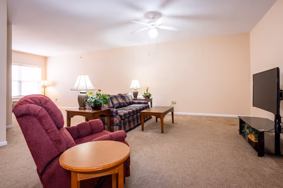 A cozy living room with beige walls and carpet. The room features a maroon recliner chair with a round wooden side table, a plaid sofa with two table lamps on wooden side tables, a rectangular wooden coffee table, and a flat-screen TV mounted on a black TV stand. There are two windows with blinds and a ceiling fan with a light fixture.