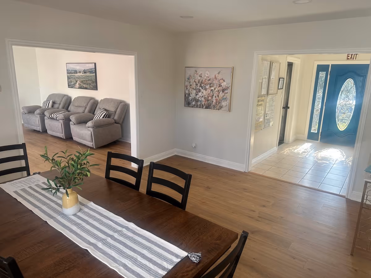 Interior view of a senior living facility showing a dining area with a wooden table and black chairs. A striped table runner and a small vase with green foliage are on the table. Beyond the dining area, there is a living room with three gray recliner chairs and a landscape painting on the wall. To the right, a hallway leads to a blue front door with decorative glass panels and an exit sign above it. The floors are wood and tile, and the walls are painted white with framed artwork and notices.