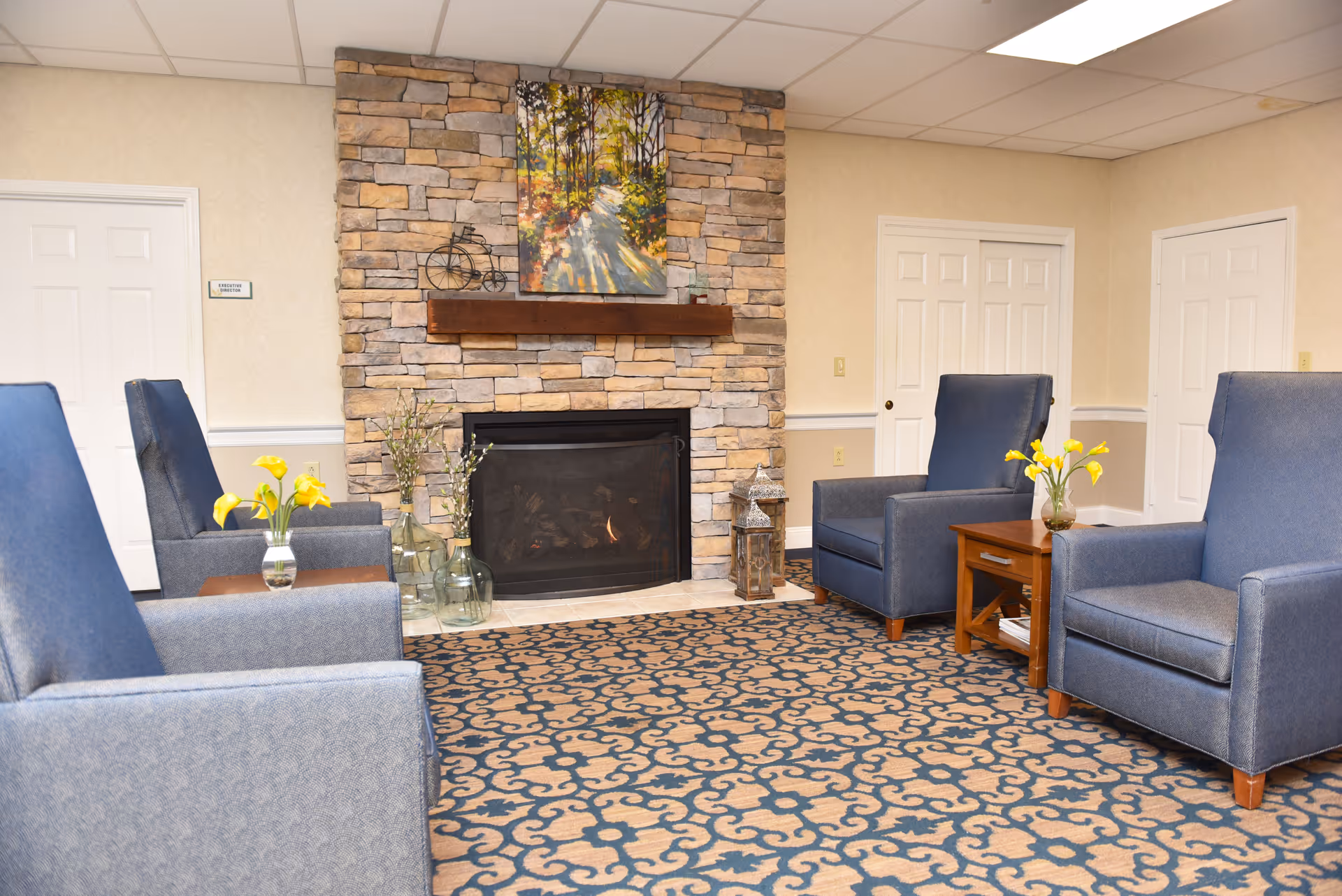 A cozy sitting area with four blue armchairs arranged around a stone fireplace. The fireplace has a wooden mantel with decorative items and a painting of a sunlit forest path above it. There are small wooden side tables with vases of yellow flowers next to the chairs, and the floor is covered with a patterned carpet. The walls are light-colored with white trim, and there are several white doors in the background.