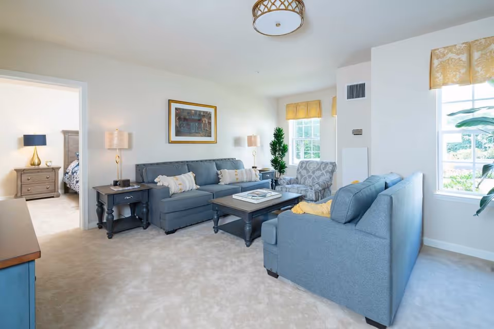 A bright and spacious living room with two blue sofas, a patterned armchair, a wooden coffee table, and two side tables with lamps. The room has beige carpet, white walls, and two large windows with yellow valances letting in natural light. A framed picture hangs on the wall above one sofa. A doorway leads to a bedroom with a nightstand and lamp visible.