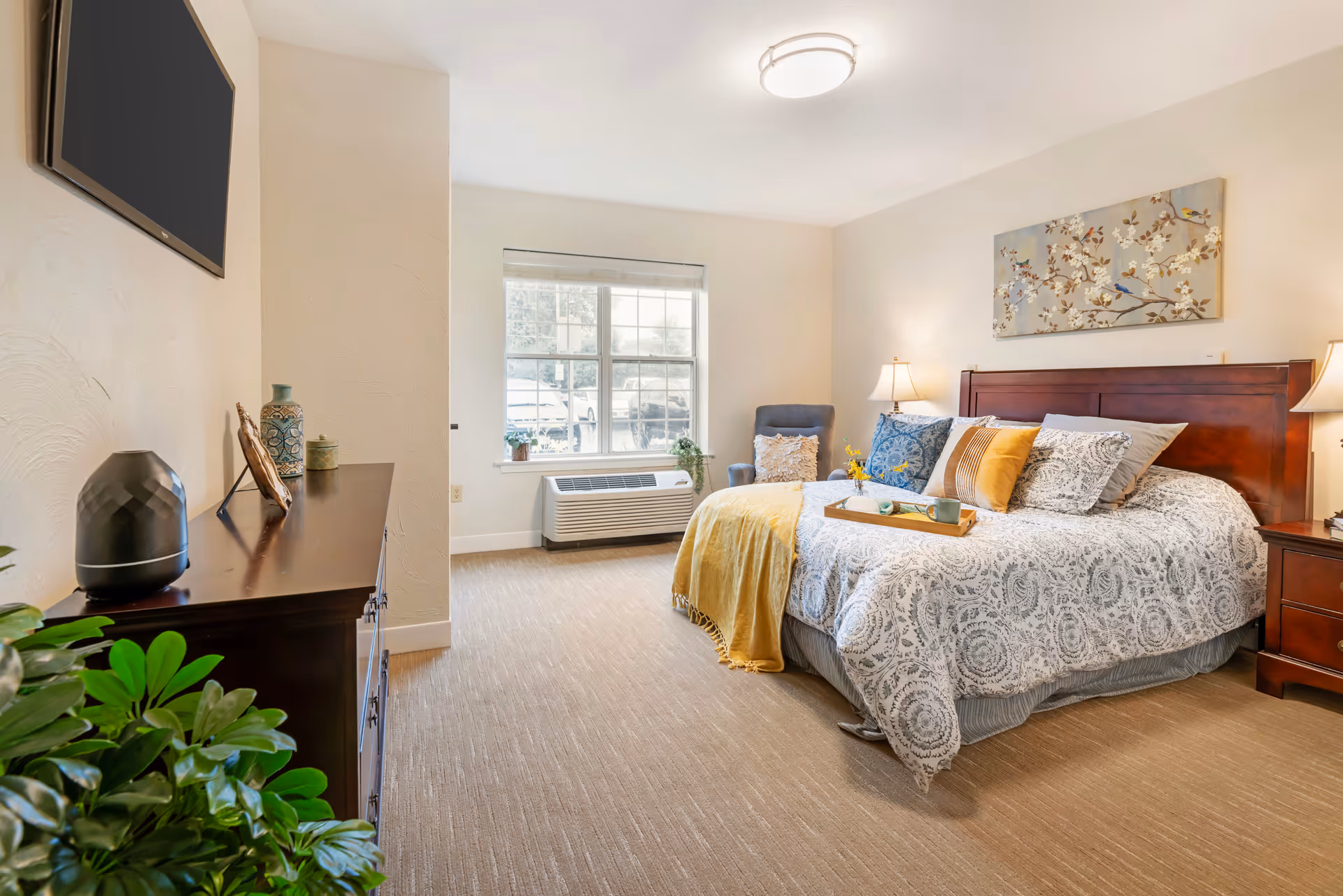 Bright, furnished bedroom with a made bed, bedside tables and lamps, a dresser with a TV, and a window with an AC unit.