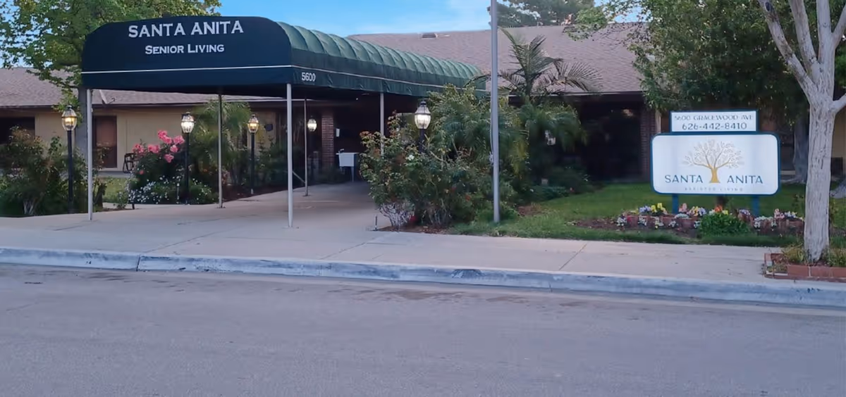 Exterior view of Santa Anita Senior Living facility entrance with a green awning, lamp posts, and landscaped garden area including flowers and bushes. A sign with the facility's name, address, and phone number is visible on the right side.
