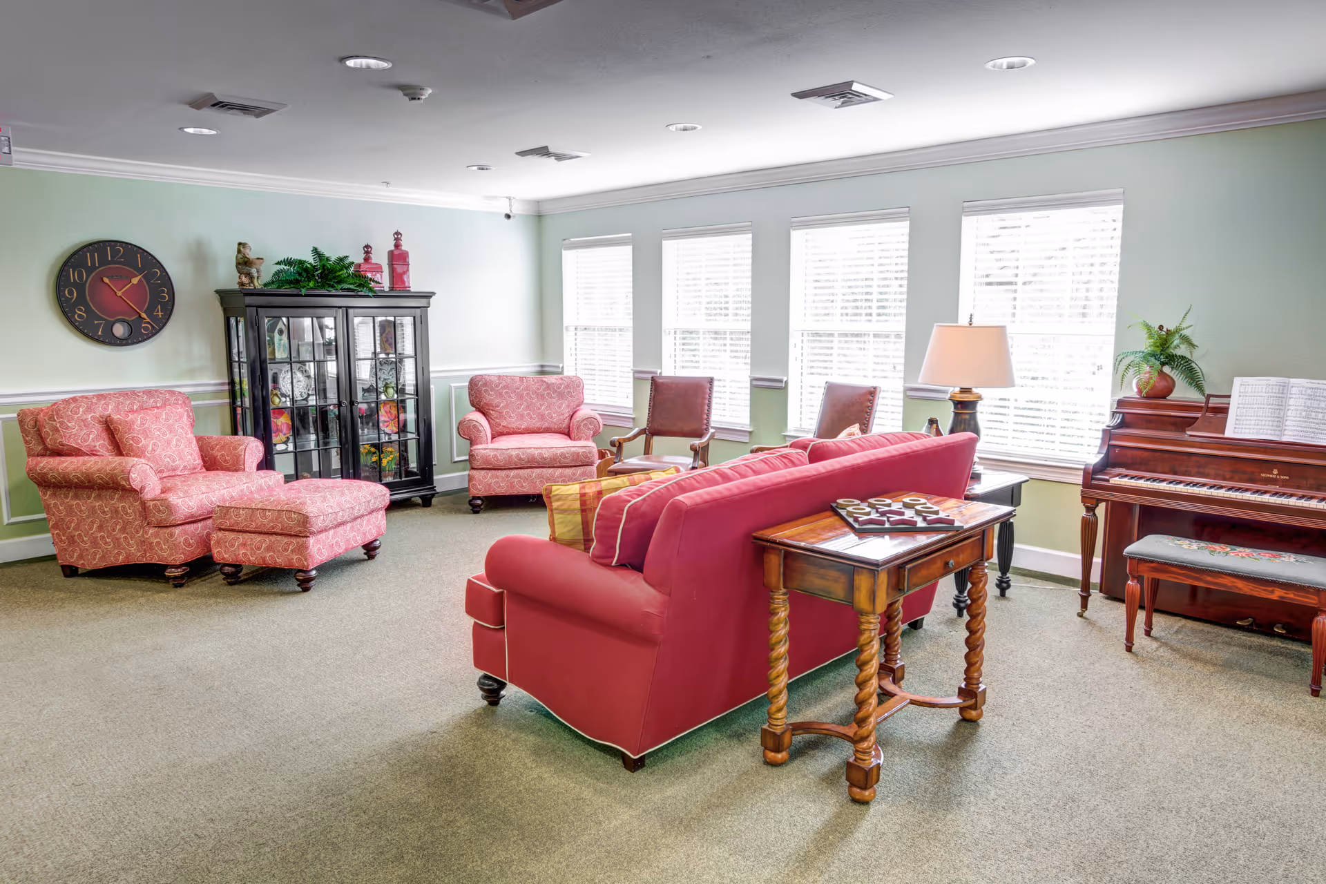 Bright common room with pink sofas and armchairs, a piano, and a glass display cabinet under windows.