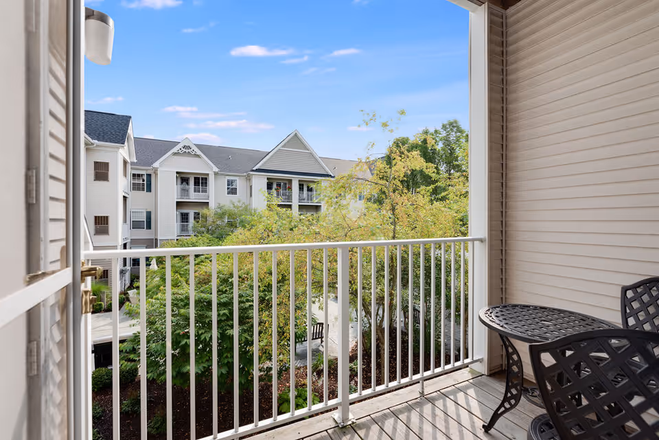 View from a balcony with white railing overlooking trees and a multi-story residential building with balconies under a blue sky.