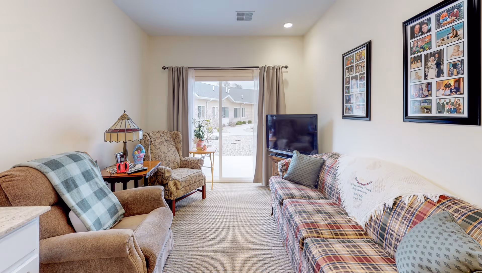 A cozy living room with a plaid sofa adorned with a white throw and green pillows, a brown armchair with a green plaid blanket, and a patterned armchair next to a small wooden side table with a decorative lamp and basket. A flat-screen TV is placed on a stand in the corner. The room has beige walls, carpeted floor, and a sliding glass door with beige curtains leading outside. Two framed photo collages hang on the wall above the sofa.