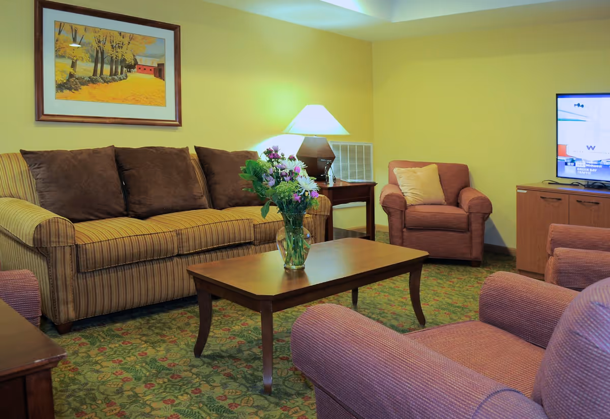 A cozy living room with a striped sofa with brown cushions, two upholstered armchairs, a wooden coffee table with a vase of flowers, a side table with a lamp, a framed painting of a fall scene on the yellow wall, and a TV on a wooden cabinet.