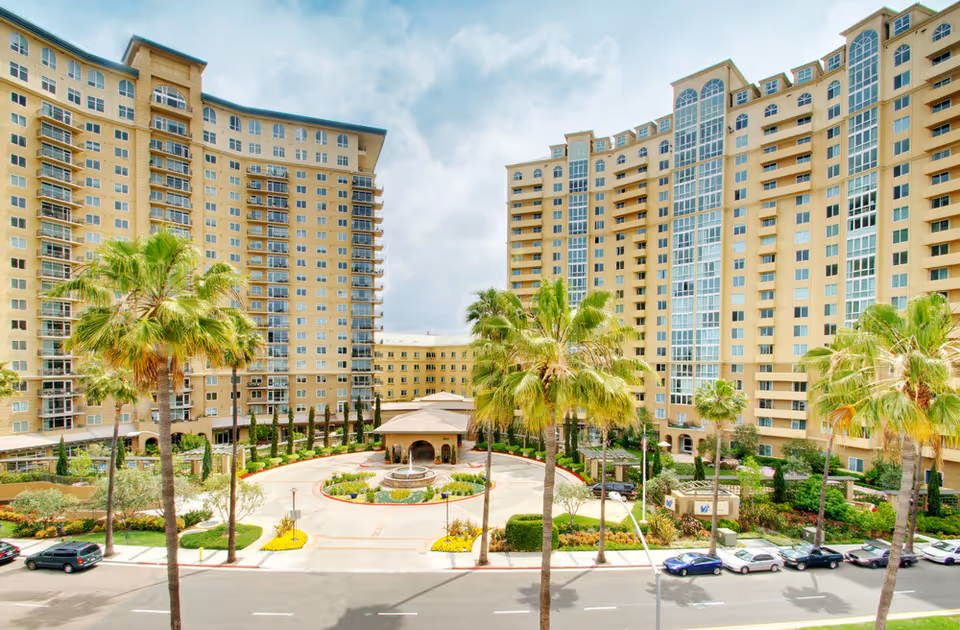 Exterior view of a large senior living facility named Vi at La Jolla Village, featuring two tall beige buildings with many windows and balconies, a circular driveway with a central fountain, palm trees, landscaped gardens, and parked cars along the street in front.