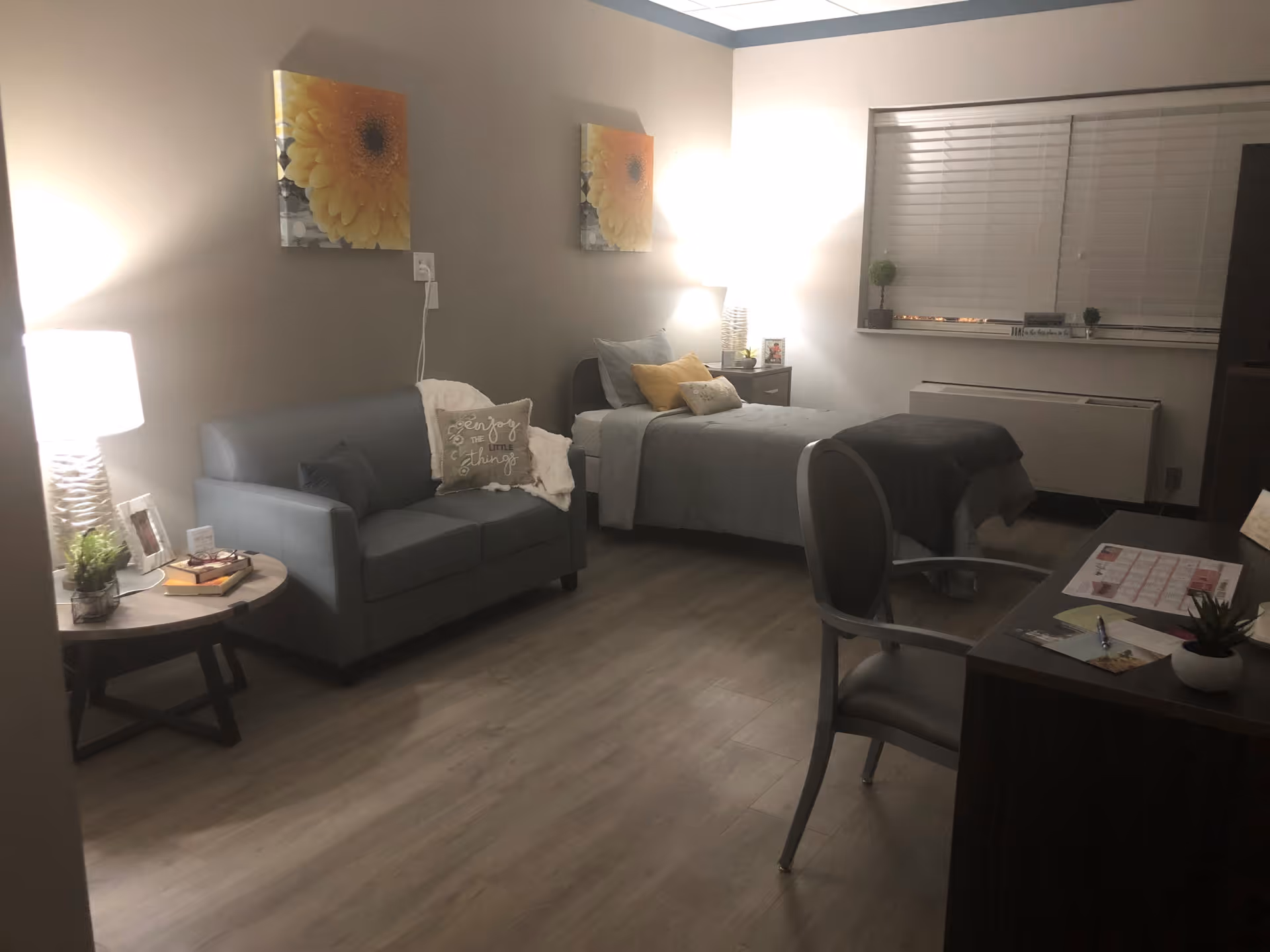 A cozy, softly lit bedroom in a senior living facility featuring a single bed with gray bedding and yellow accent pillows, a gray loveseat with decorative pillows, a round side table with a lamp and books, two yellow flower paintings on the wall, a desk with a chair, and a window with blinds.