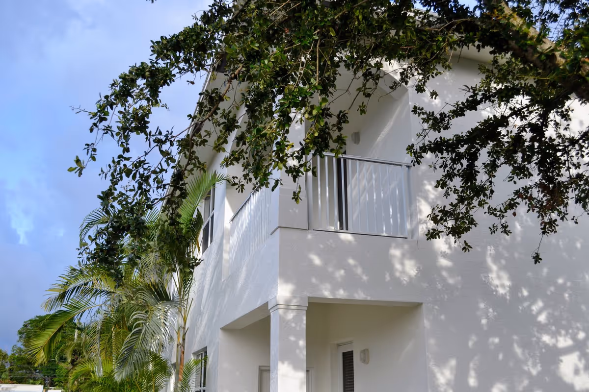 Exterior view of a white two-story building with a balcony on the upper floor, partially shaded by tree branches and surrounded by palm trees under a blue sky.