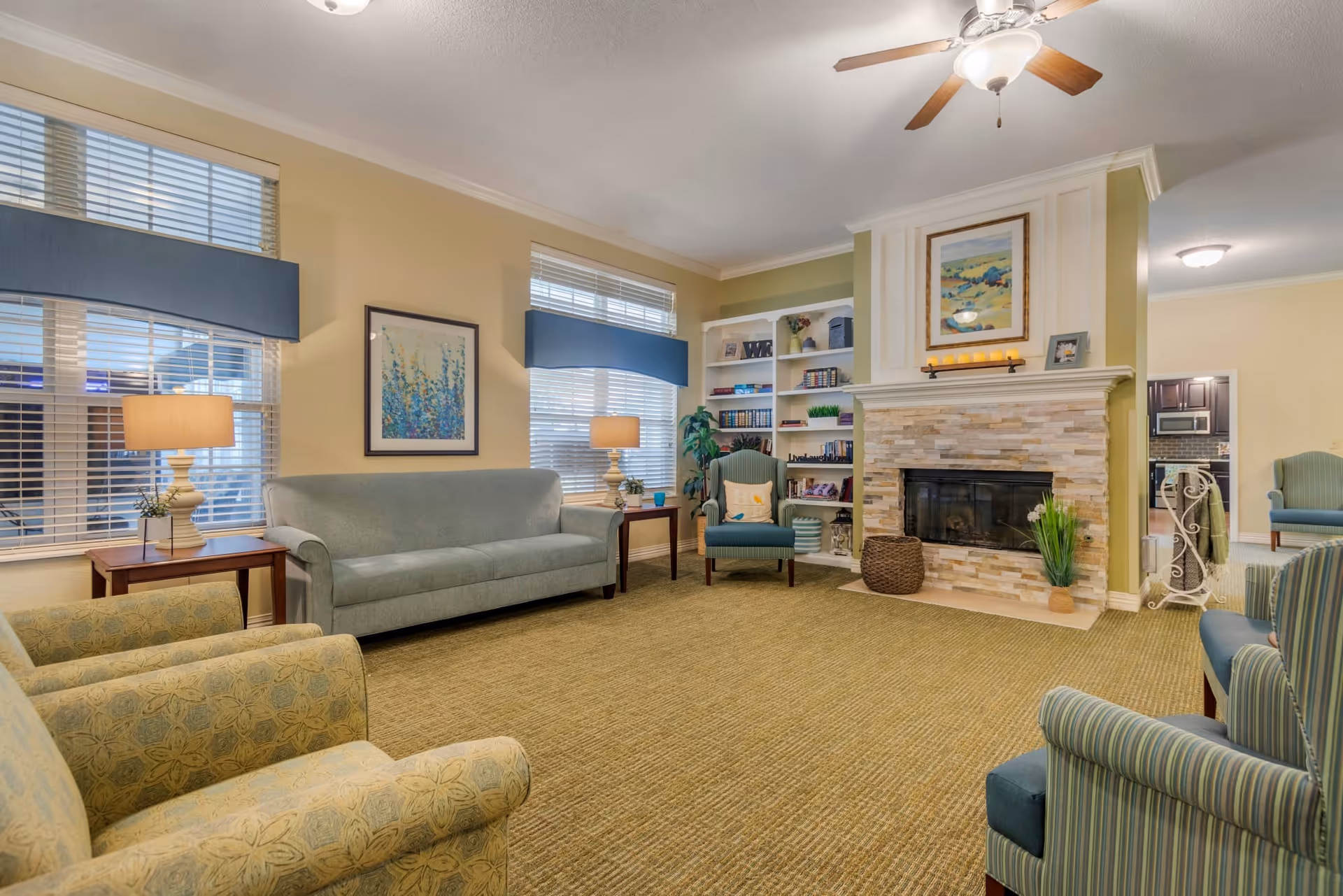 A cozy living room with a green carpet, a stone fireplace with a painting above it, built-in white shelves filled with books and decor, a green armchair with a pillow, a blue sofa, two side tables with lamps, and multiple windows with blue valances allowing natural light.