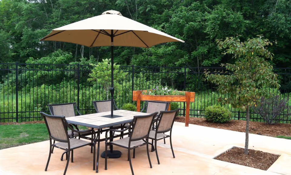Outdoor patio with a table, six chairs and a large umbrella beside a fenced wooded area.