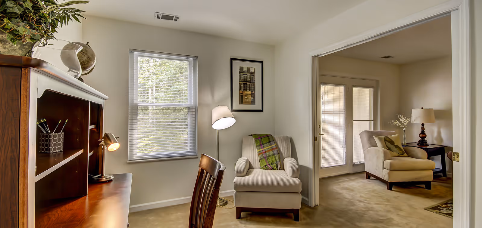 Cozy interior sitting area with upholstered armchairs, a wooden desk, floor lamp, window blinds and French doors to an adjoining room.