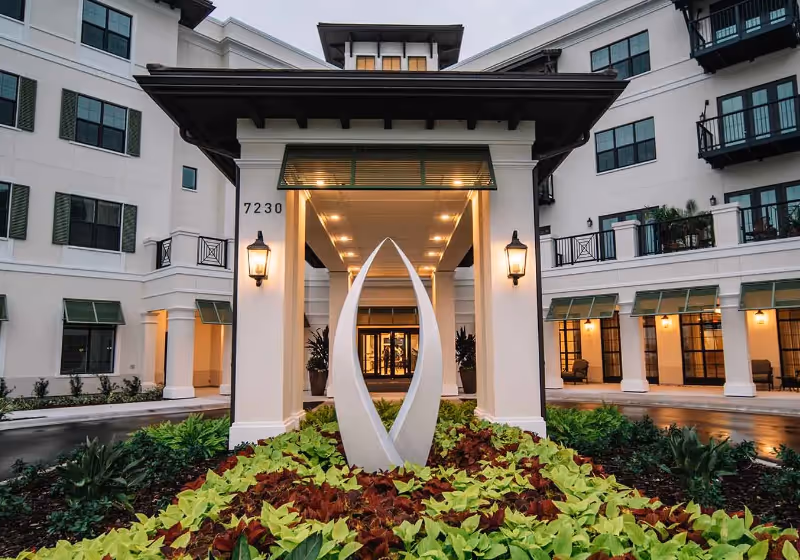 Entrance of a senior living facility named Grand Living At Lakewood Ranch, featuring a covered driveway with two lantern-style lights on columns, a modern white sculpture in the landscaped garden bed, and a multi-story building with green window awnings and balconies in the background during dusk.