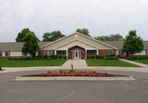 Front exterior of a single-story memory care building with a central entrance, circular driveway, lawn, trees, and a flowerbed.