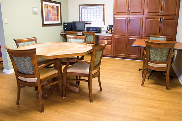 A spacious dining room featuring an octagonal wooden table surrounded by six chairs, with a computer station and wooden cabinets in the background.