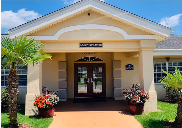 Front entrance of The Waterford at Creekside administration building with double glass doors, beige exterior walls, a small palm tree and potted plants with red flowers on either side of the entrance, and a sign above the door that reads 'ADMINISTRATION'.