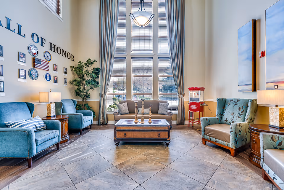 Bright senior living common room with tall windows, blue armchairs, a sofa, coffee table and a 'Wall of Honor' display.
