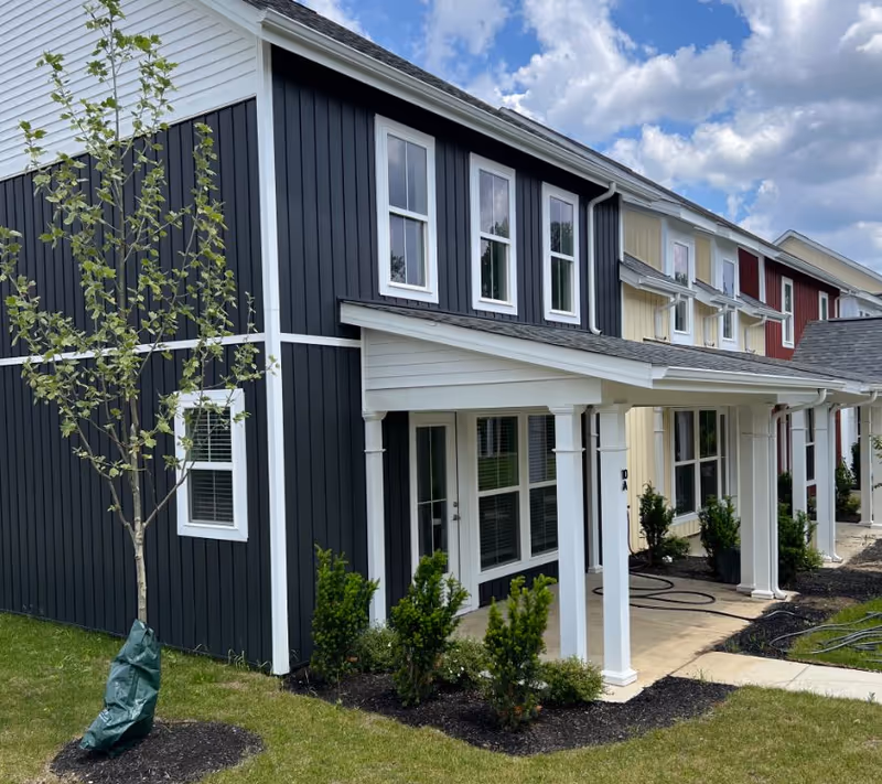 Exterior view of a modern senior living facility building with multiple units. The building features vertical siding in dark gray, cream, and red colors with white trim. Each unit has large windows and a covered porch supported by white columns. There are small landscaped bushes and a young tree planted in front of the building, with a partly cloudy sky in the background.