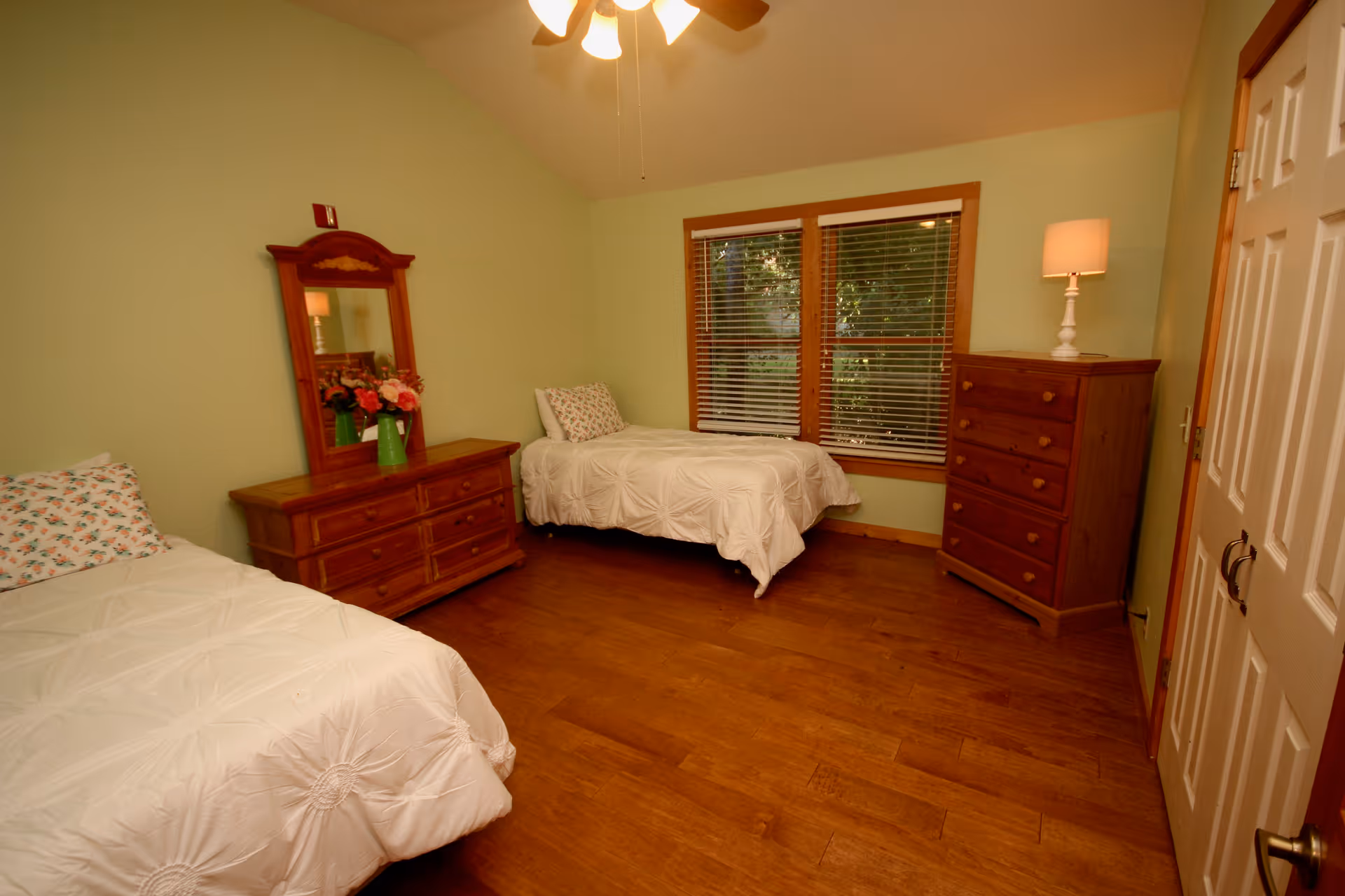 A bedroom with two single beds, each with white bedding and floral pillows. The room has light green walls, wooden flooring, a wooden dresser with a mirror and flowers on top, a wooden chest of drawers with a lamp, and a window with blinds showing greenery outside. A ceiling fan with lights is visible above.