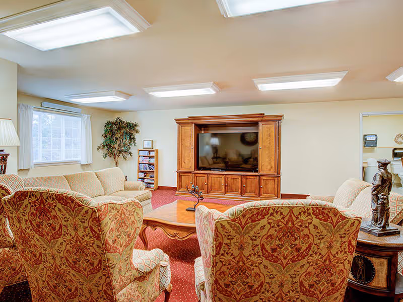 A cozy living room in an assisted living facility featuring patterned armchairs and a beige sofa arranged around a wooden coffee table. A large wooden entertainment center with a flat-screen TV is against the far wall. The room has a red carpet, beige walls, a window with white curtains, and ceiling lights. There is a small bookshelf and a decorative plant near the window.