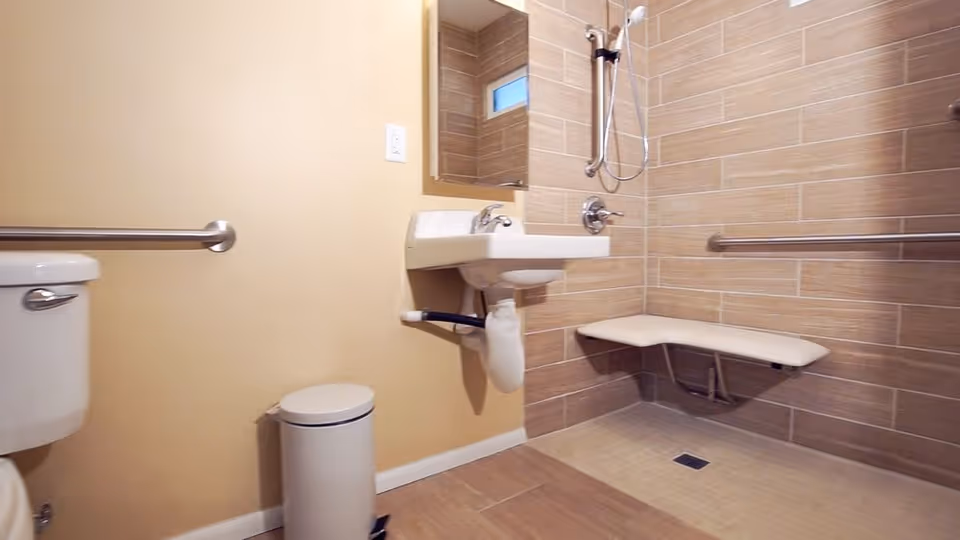 Accessible bathroom with grab bars, a wall-mounted sink, toilet, and a folding shower bench.
