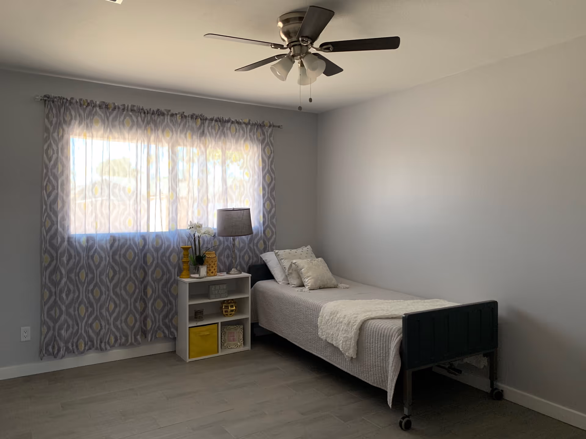 A simple bedroom with a single bed against the right wall, covered with a light gray bedspread and several pillows. Next to the bed is a small white shelving unit with decorative items including a lamp, a plant, and yellow accents. A large window with patterned curtains lets in natural light, and a ceiling fan with lights is mounted on the ceiling.