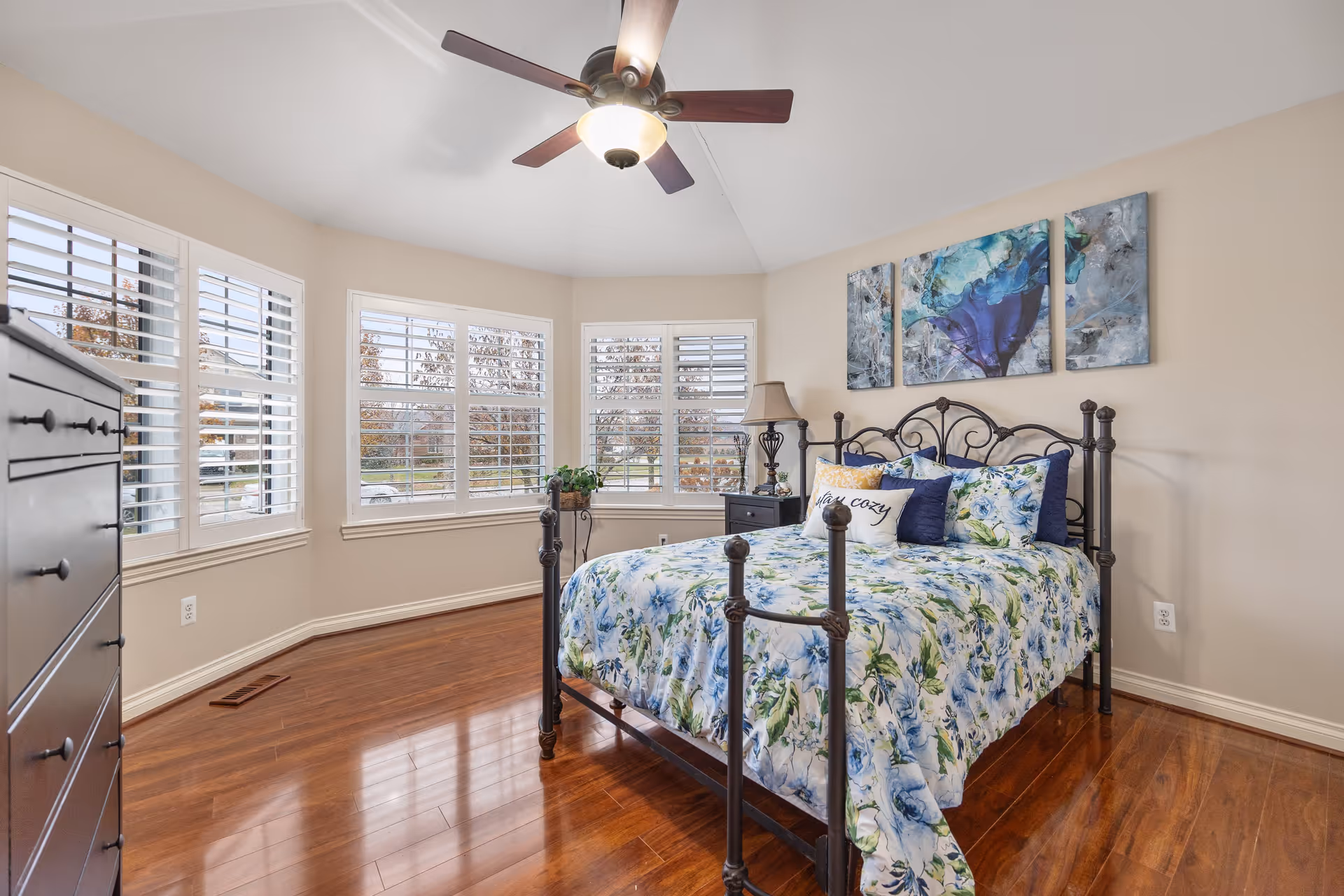 A bright bedroom with a metal framed bed adorned with floral bedding and multiple pillows, including one that says 'stay cozy'. The room features large windows with white plantation shutters, a ceiling fan with a light, a black dresser, a nightstand with a lamp, and three abstract blue and green paintings on the wall above the bed. The floor is polished wood.