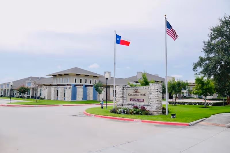 Exterior view of Orchard Park at Southfork Assisted & Senior Living facility with a stone sign displaying the facility name, two flagpoles with the Texas state flag and the American flag, and a well-maintained lawn and trees surrounding the building.