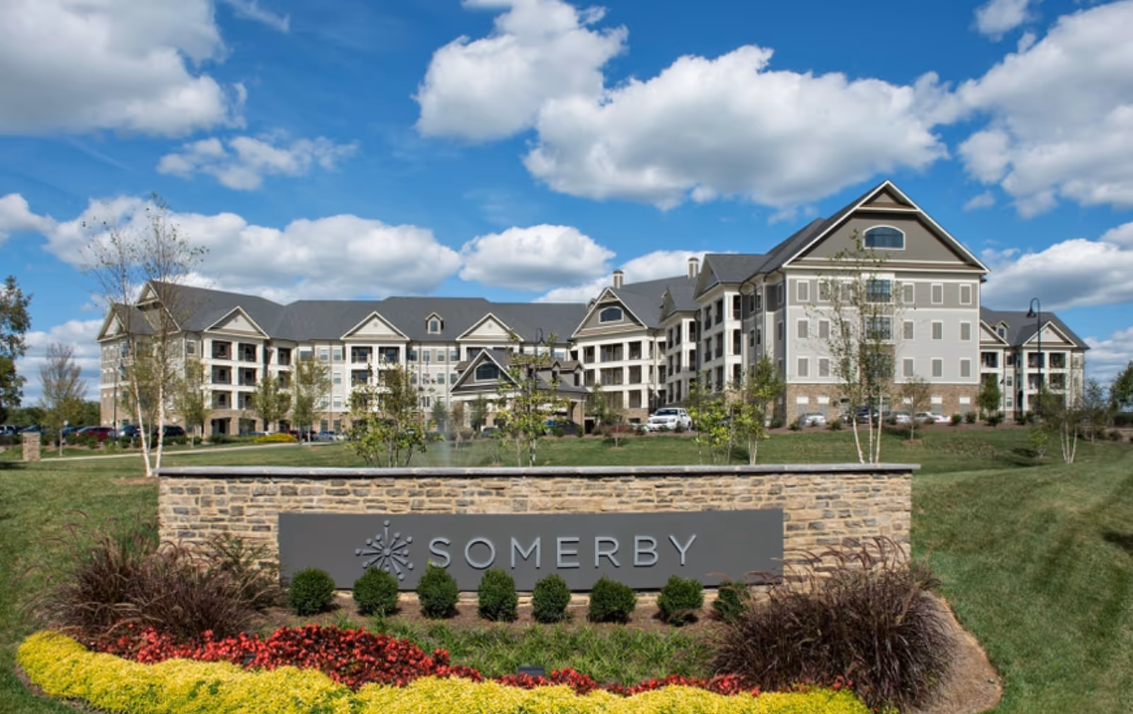 Exterior view of the Somerby Franklin senior living facility building under a blue sky with scattered clouds. The building is large with multiple floors, beige siding, and a gray roof. In the foreground, there is a stone sign with the word 'SOMERBY' and landscaped greenery including bushes and colorful flowers.