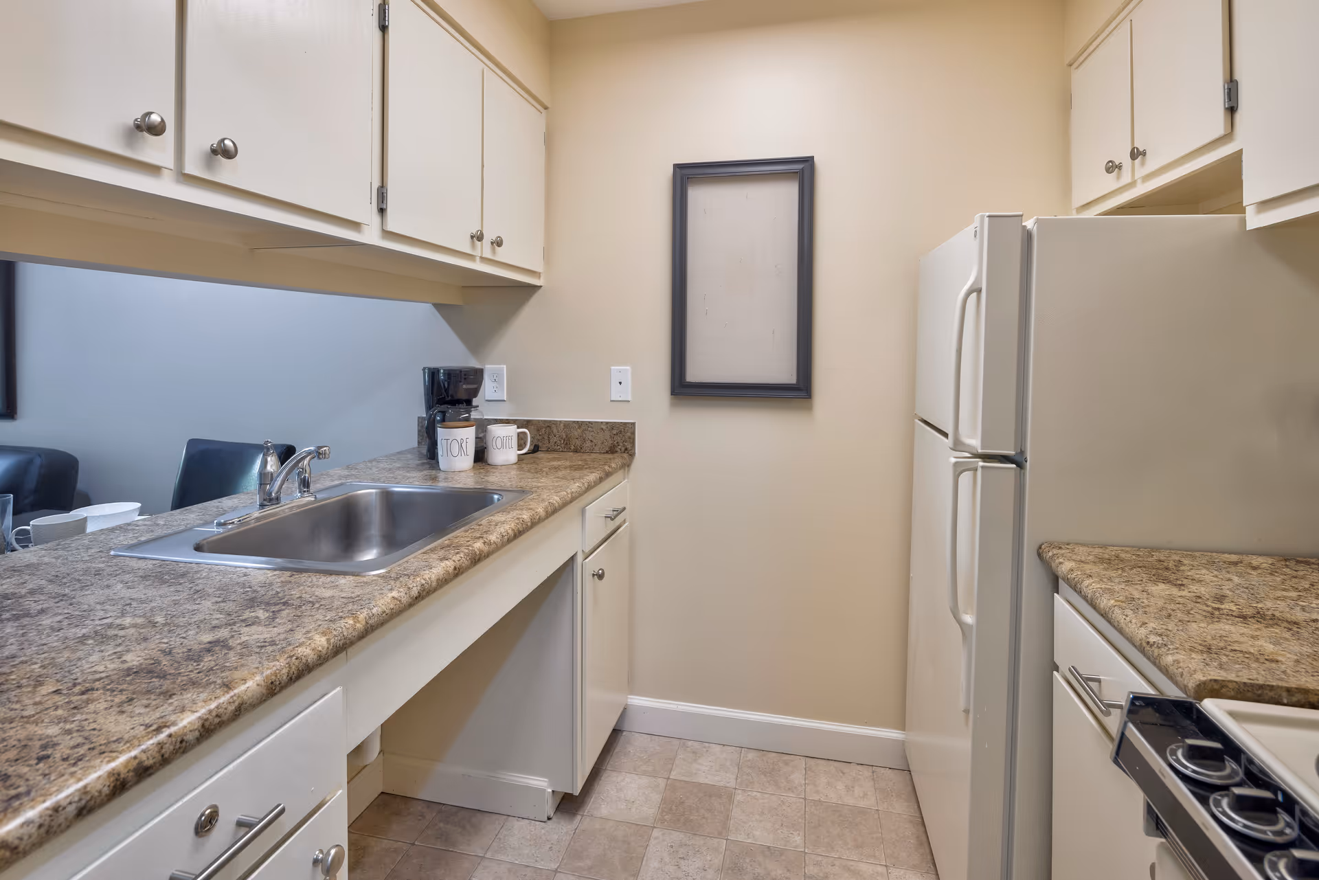 A small kitchen area with beige walls and tiled floor, featuring a countertop with a stainless steel sink, a coffee maker, and two mugs. There are white cabinets above and below the countertop, a white refrigerator, and a stove with control knobs visible. A framed empty canvas or bulletin board hangs on the wall.