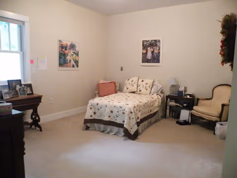 A simple bedroom with a single bed covered in a floral quilt, two framed pictures on the wall, a small bedside table with a lamp and some items, a cushioned armchair, and a window letting in natural light.