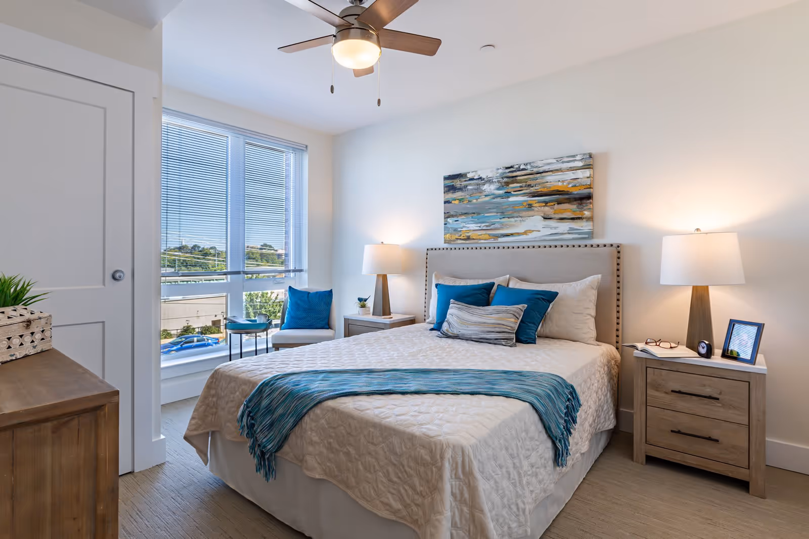 A bright and neatly arranged bedroom with a large bed covered in a white quilt and blue accent pillows and throw. There are two wooden nightstands on either side of the bed, each with a lamp. A modern ceiling fan with a light hangs above. A large window with blinds lets in natural light, and a chair with a blue pillow and a small side table are placed near the window. A colorful abstract painting hangs above the bed.