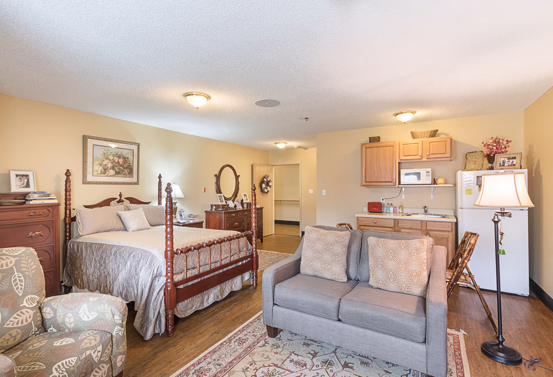 A furnished senior living suite with bed, sofa, kitchenette, and dresser in warm tones.