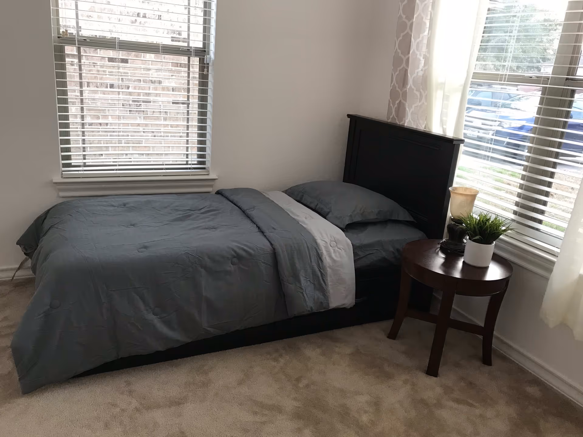 A small bedroom with a single bed made with gray bedding and a dark wooden headboard. Next to the bed is a round wooden side table with a small potted plant and a decorative item. The room has beige carpet and two windows with white blinds, one behind the bed and one to the right, letting in natural light.