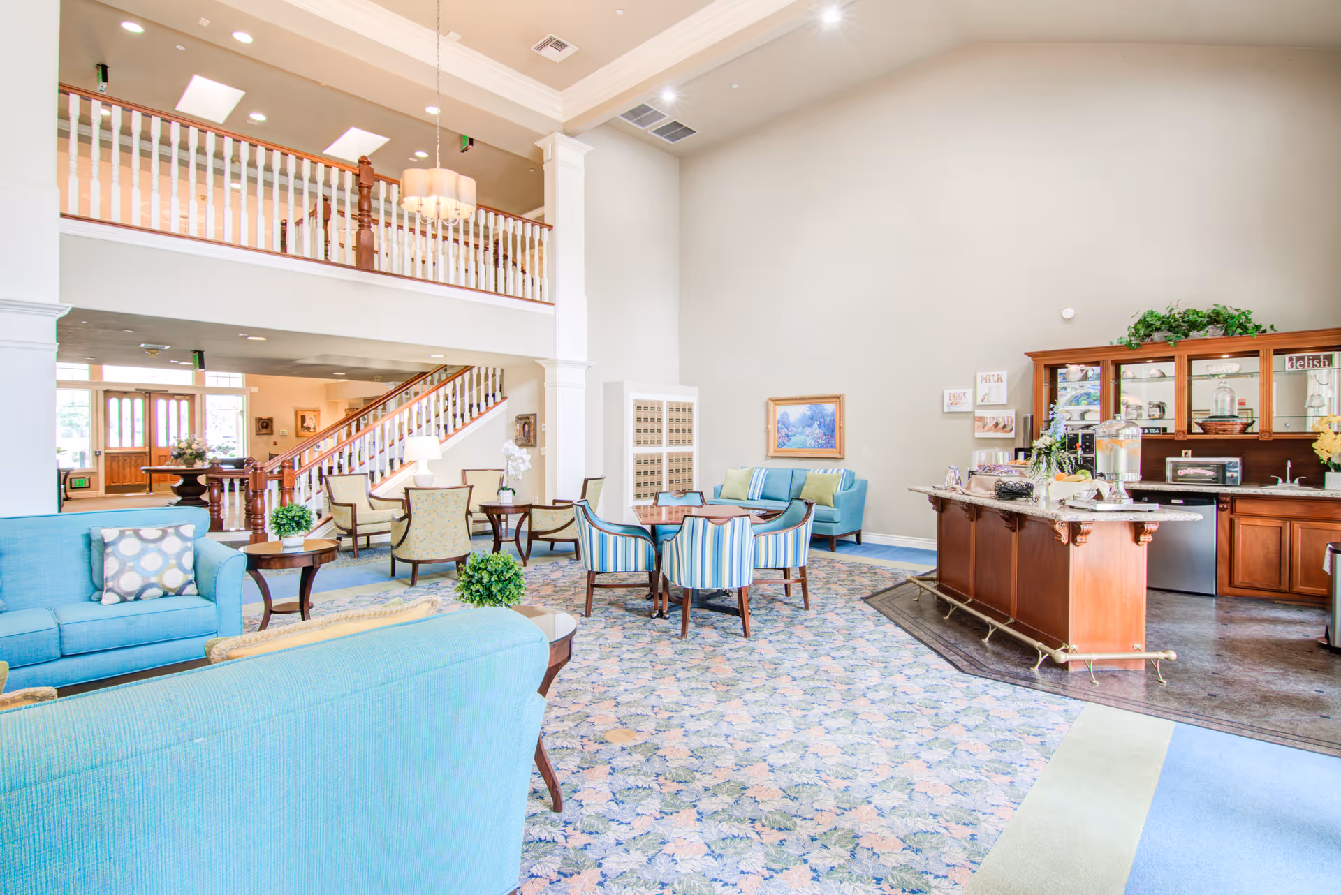 Bright and spacious senior living common area with high ceilings, light blue sofas, patterned carpet, wooden tables and chairs, a wooden kitchen island with refreshments, and a staircase leading to an upper level with white railings.