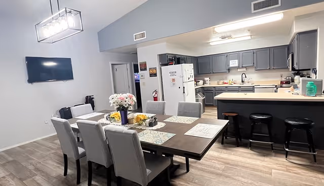 Open dining area with a large table and upholstered chairs adjacent to an open kitchen with gray cabinets and bar stools.