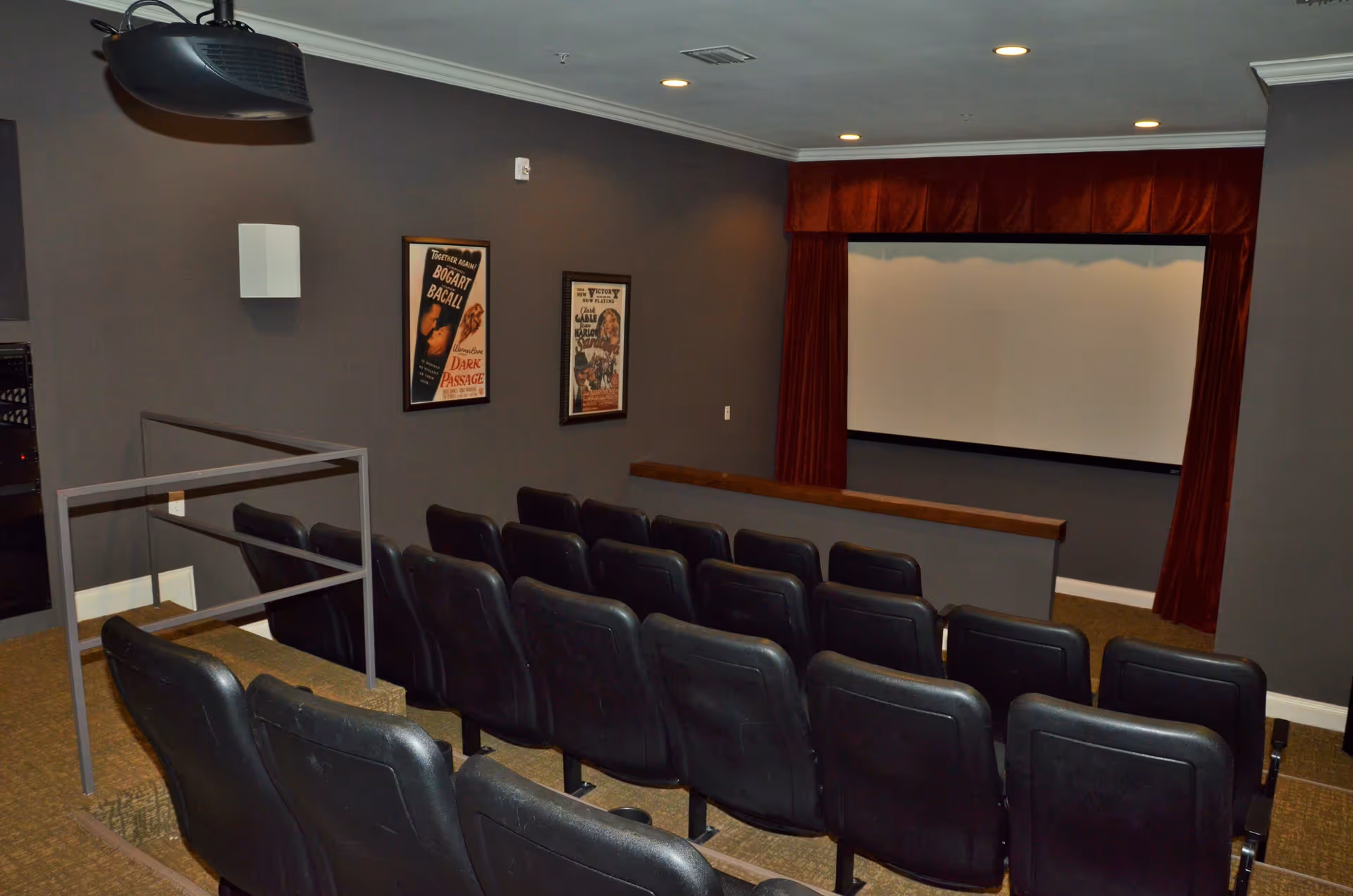 Small movie theater room with three rows of black cushioned seats facing a large white projection screen framed by red curtains. The walls are painted gray and decorated with vintage movie posters. Ceiling lights illuminate the room.