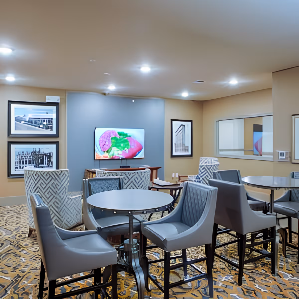A cozy common area with multiple round tables surrounded by cushioned chairs. The room has patterned carpet flooring, beige walls, and recessed ceiling lights. A flat-screen TV is mounted on a blue accent wall, displaying a colorful image. Framed black and white photographs hang on the walls, and a large mirror reflects part of the room.