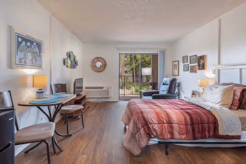 A cozy bedroom in a senior living facility with a bed covered in red and beige bedding, a small round table with two chairs, a TV on a stand, a recliner chair near a sliding glass door leading to a balcony, and various framed pictures and decorations on the walls.