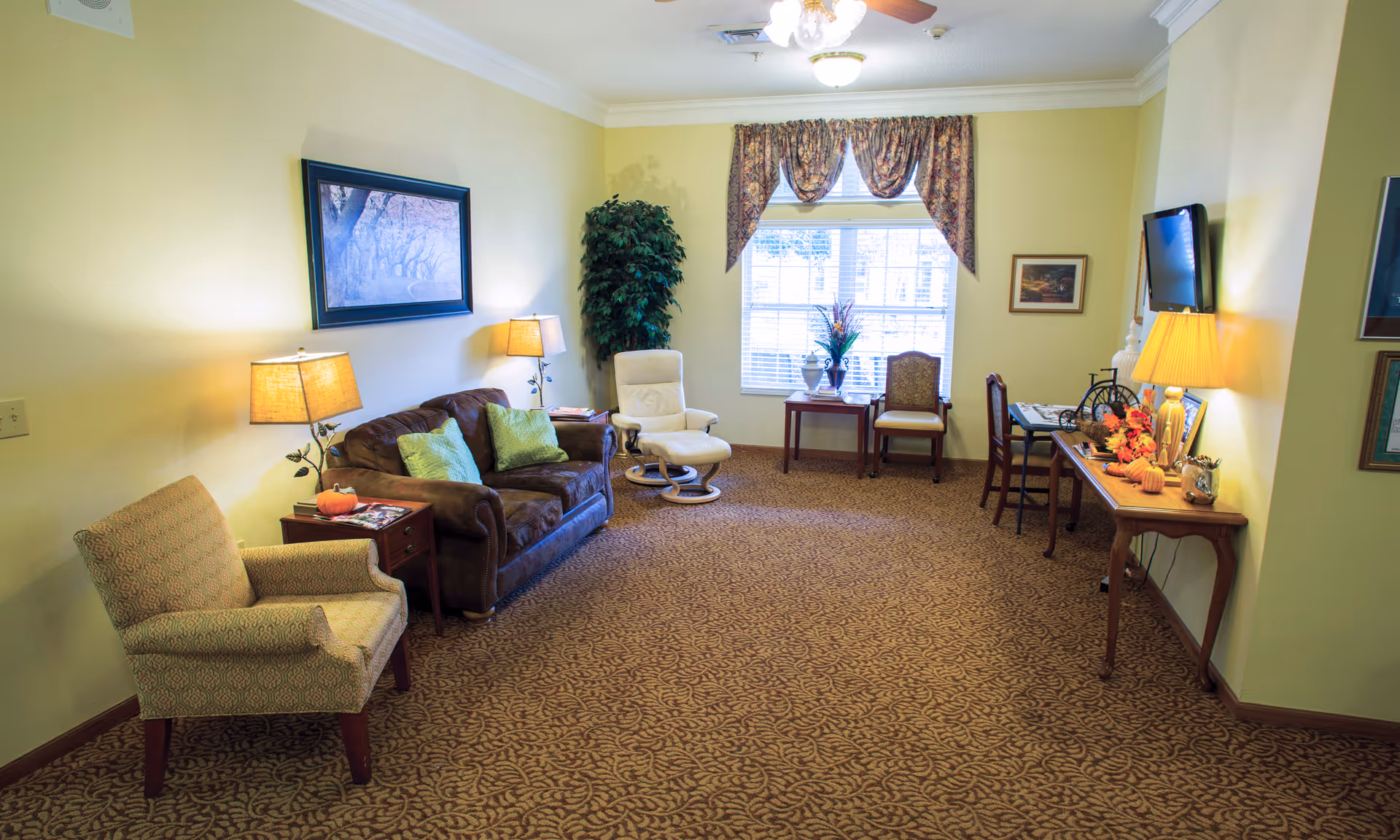 A cozy living room with yellow walls and patterned carpet. The room features a brown sofa with green pillows, a beige armchair, a white recliner, and a small wooden table with two chairs near a window with floral curtains. There are two table lamps, a wall-mounted TV, framed pictures on the walls, and decorative items on a console table.