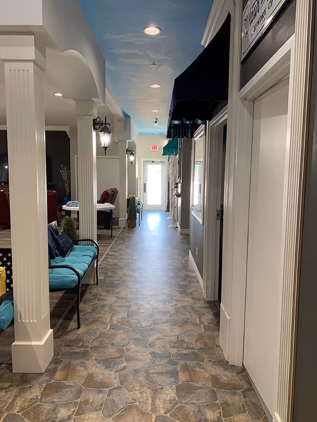 A hallway in a senior living facility with stone-patterned flooring, white columns, and wall-mounted lantern-style lights. There are cushioned benches along the left side and doors on the right side, with a ceiling painted to resemble a blue sky with clouds. The hallway leads to a glass door at the far end.