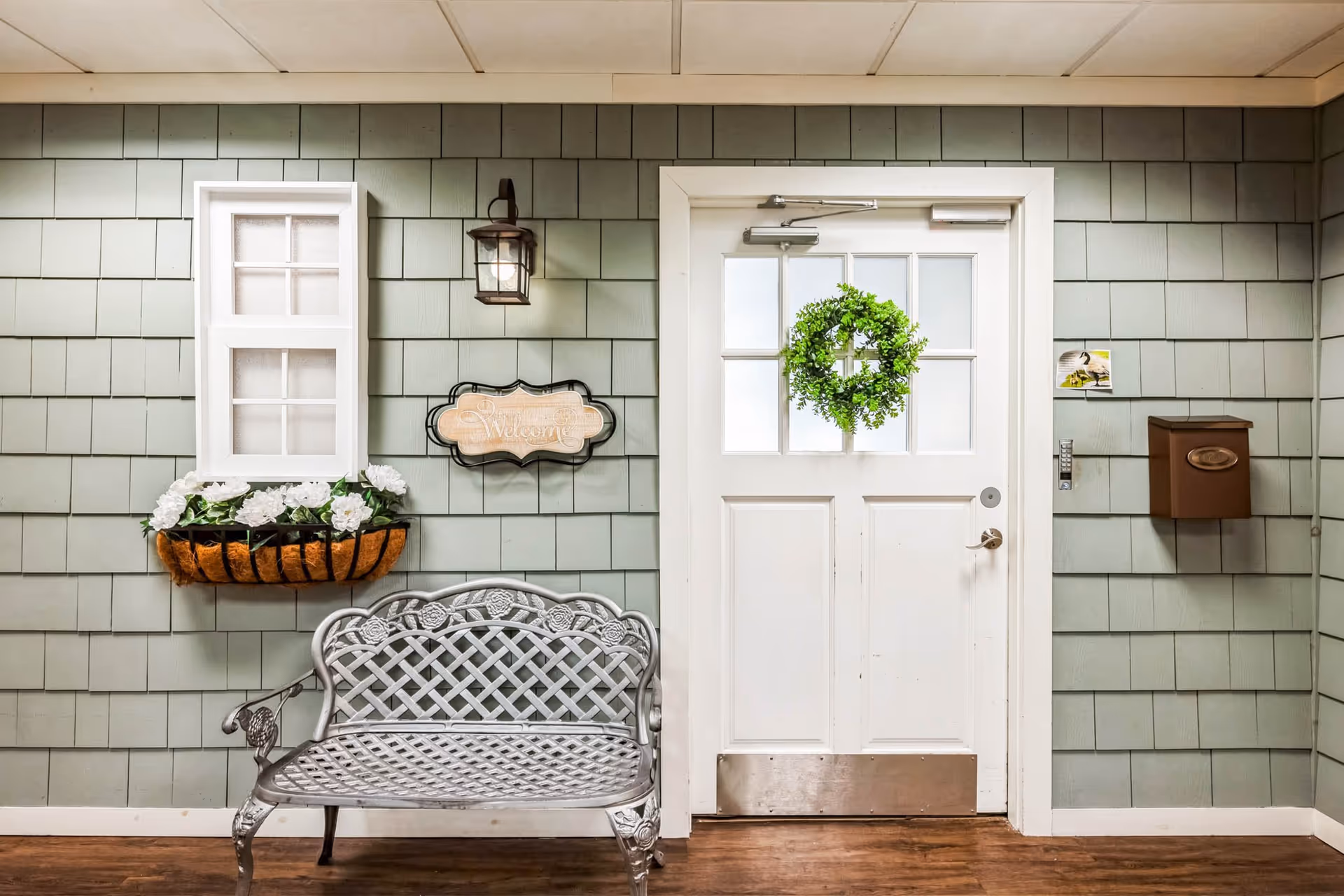 Interior view of an entryway with a white door decorated with a green wreath, a small window with white flowers in a planter box, a wall-mounted lantern light, a wooden welcome sign, a brown mailbox, and a decorative metal bench on a wooden floor.
