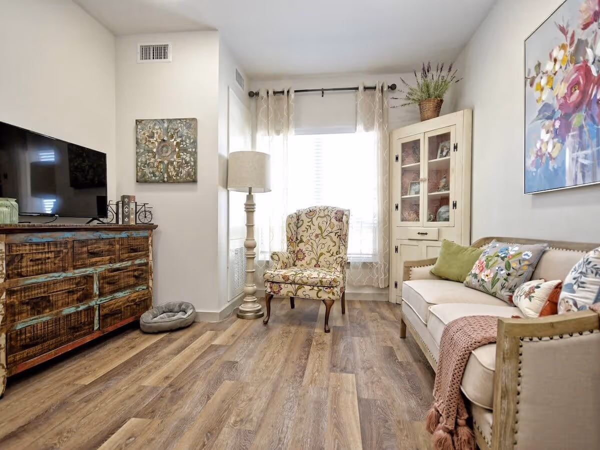 A cozy living room with a rustic wooden dresser holding a flat-screen TV on the left, a floral patterned armchair in front of a window with sheer curtains, a tall floor lamp, a cream-colored cabinet with glass doors and decorative items inside, and a beige sofa adorned with colorful floral and green pillows and a pink knitted throw blanket. The room has wood flooring and light-colored walls with floral artwork.
