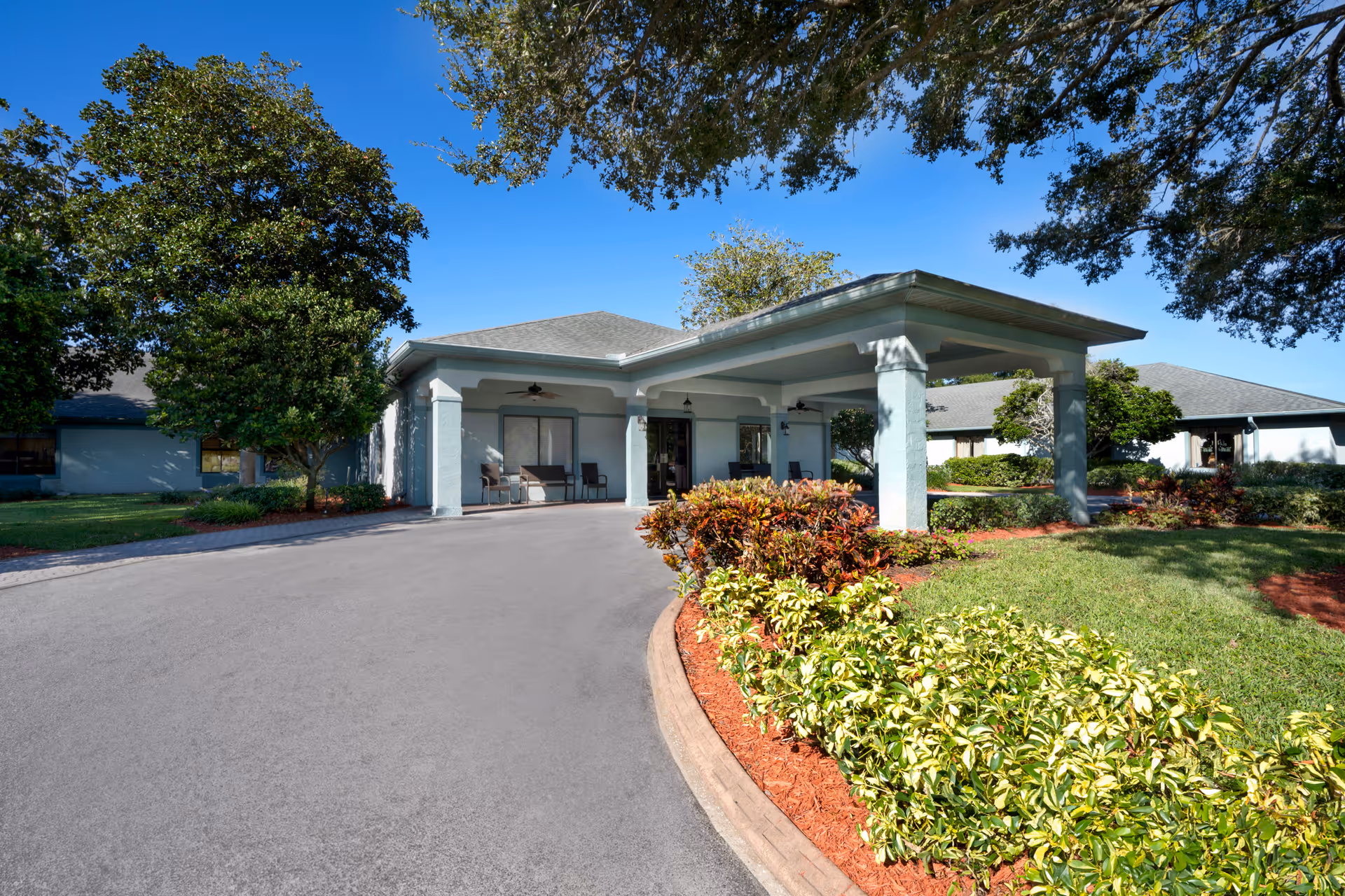 Exterior view of Bayonet Point Health Center showing the entrance with a covered drop-off area, surrounded by well-maintained landscaping including green shrubs, trees, and a clear blue sky.