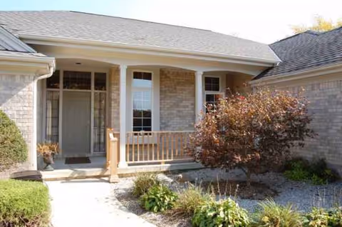 Exterior view of a senior living facility with a front porch, entry door, and shrubs.