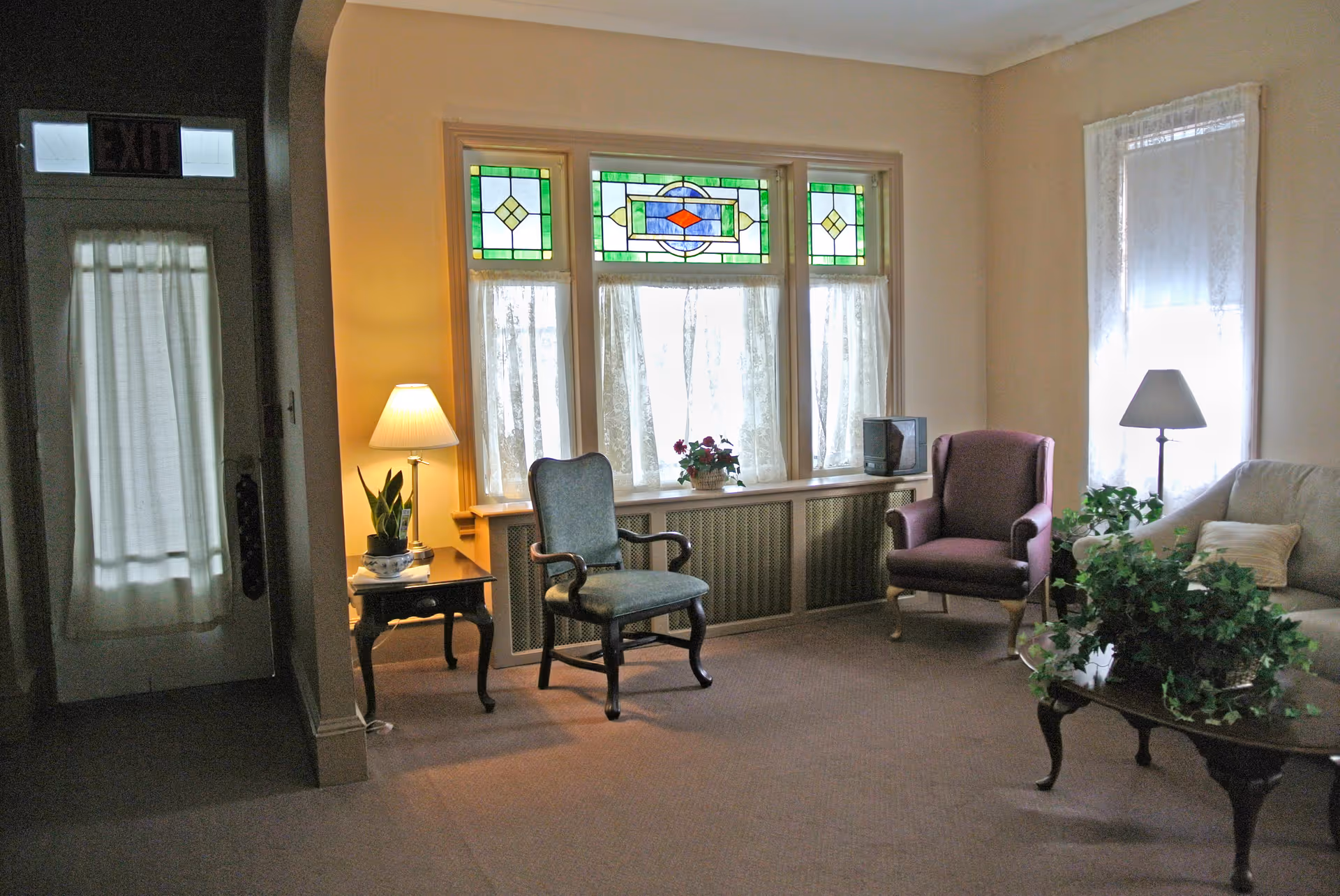 A cozy living room with beige walls and carpet, featuring a stained glass window with green, yellow, and red accents. The room contains a blue upholstered armchair, a purple armchair, a beige sofa with a pillow, a wooden side table with a lamp and a potted plant, and a wooden coffee table with a large green leafy plant. There is a door with a curtain and an exit sign above it on the left side of the room.
