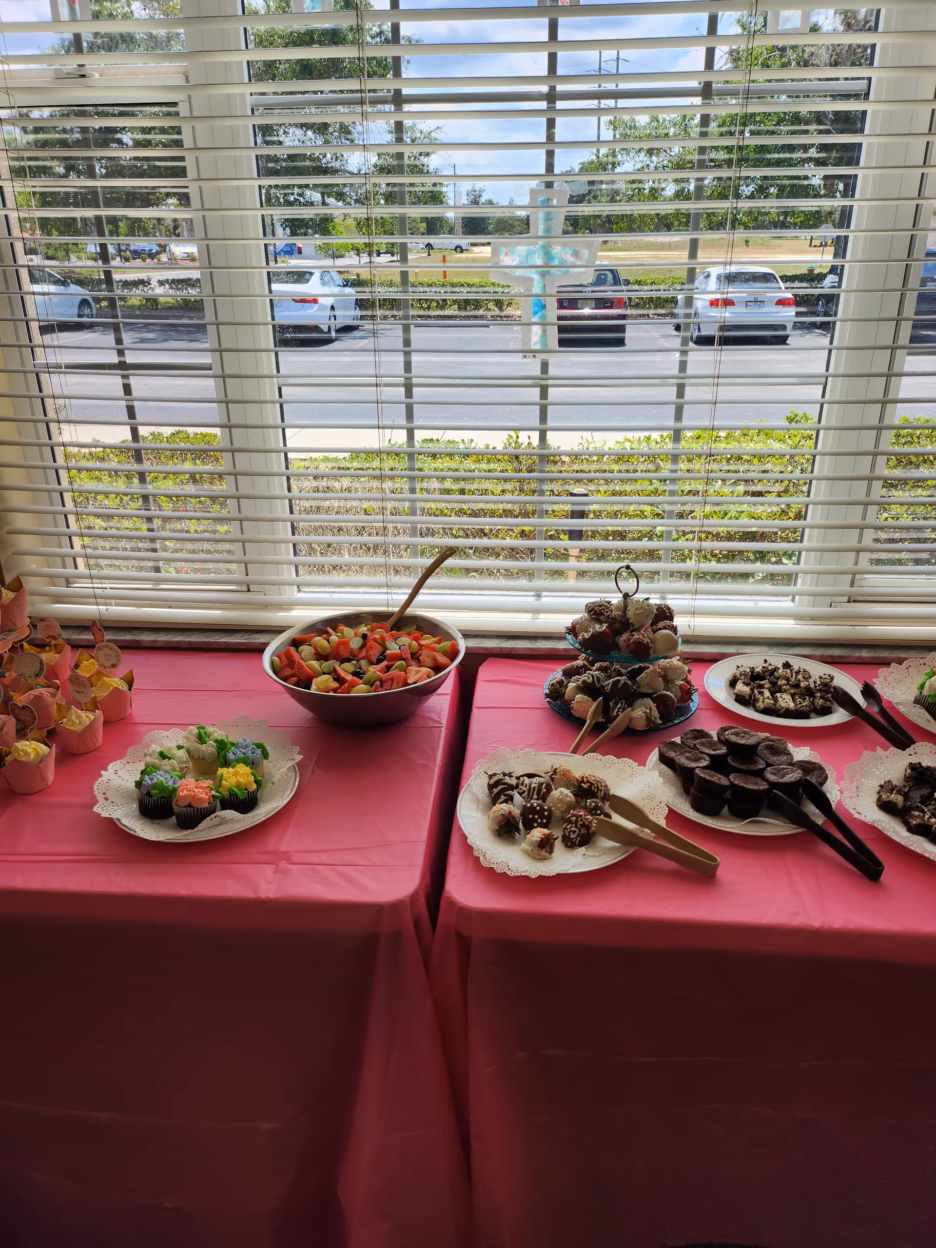 A table covered with a pink tablecloth is set up in front of a window with white blinds. On the table, there are various desserts including cupcakes with colorful frosting, a bowl of mixed fruit salad, chocolate-covered treats on a tiered stand, and plates with brownies and other sweets. Outside the window, cars are parked along a street with greenery and trees in the background.