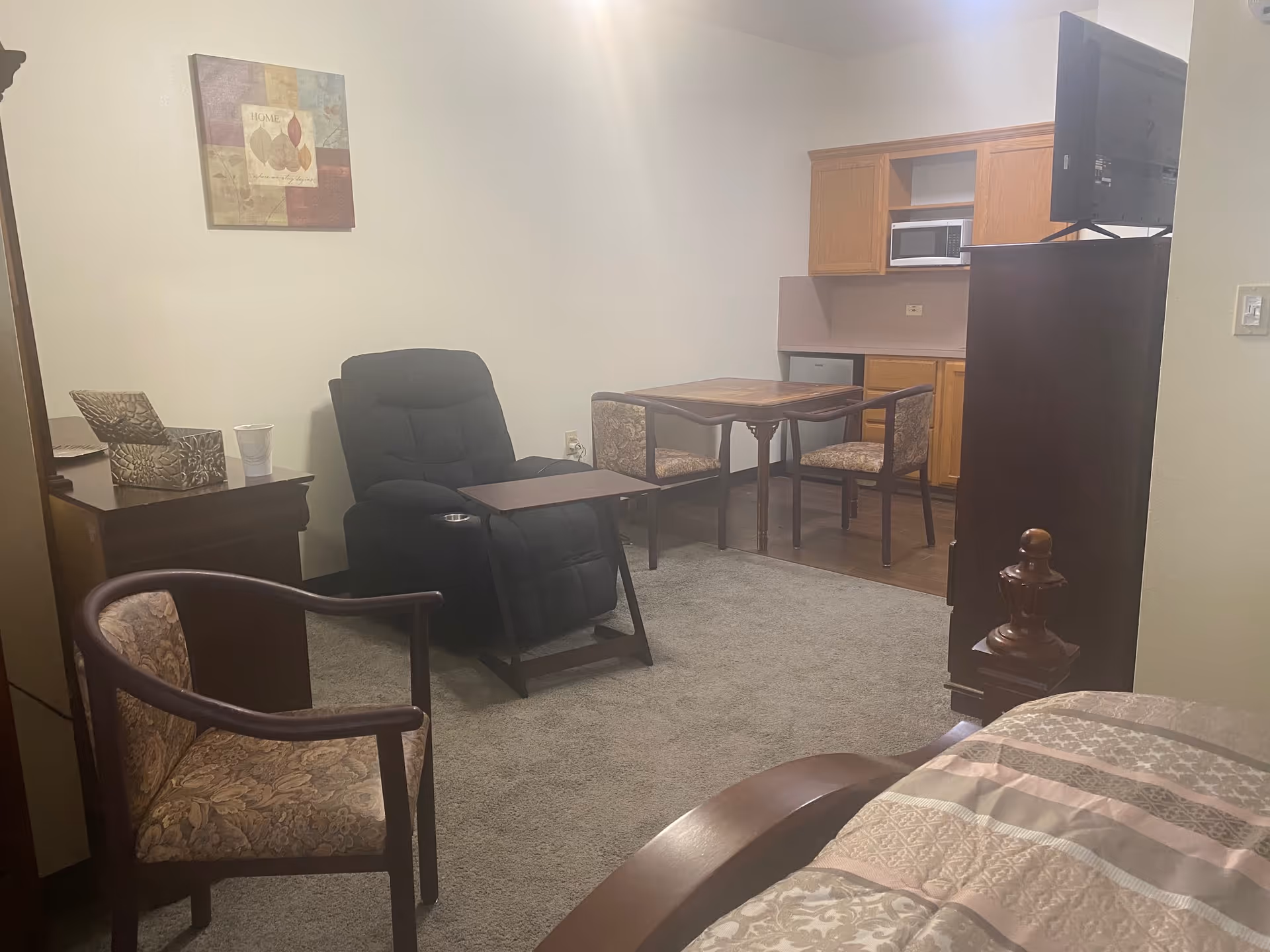 Studio living area with a recliner, dining table and chairs, kitchenette cabinets and a partially visible bed.